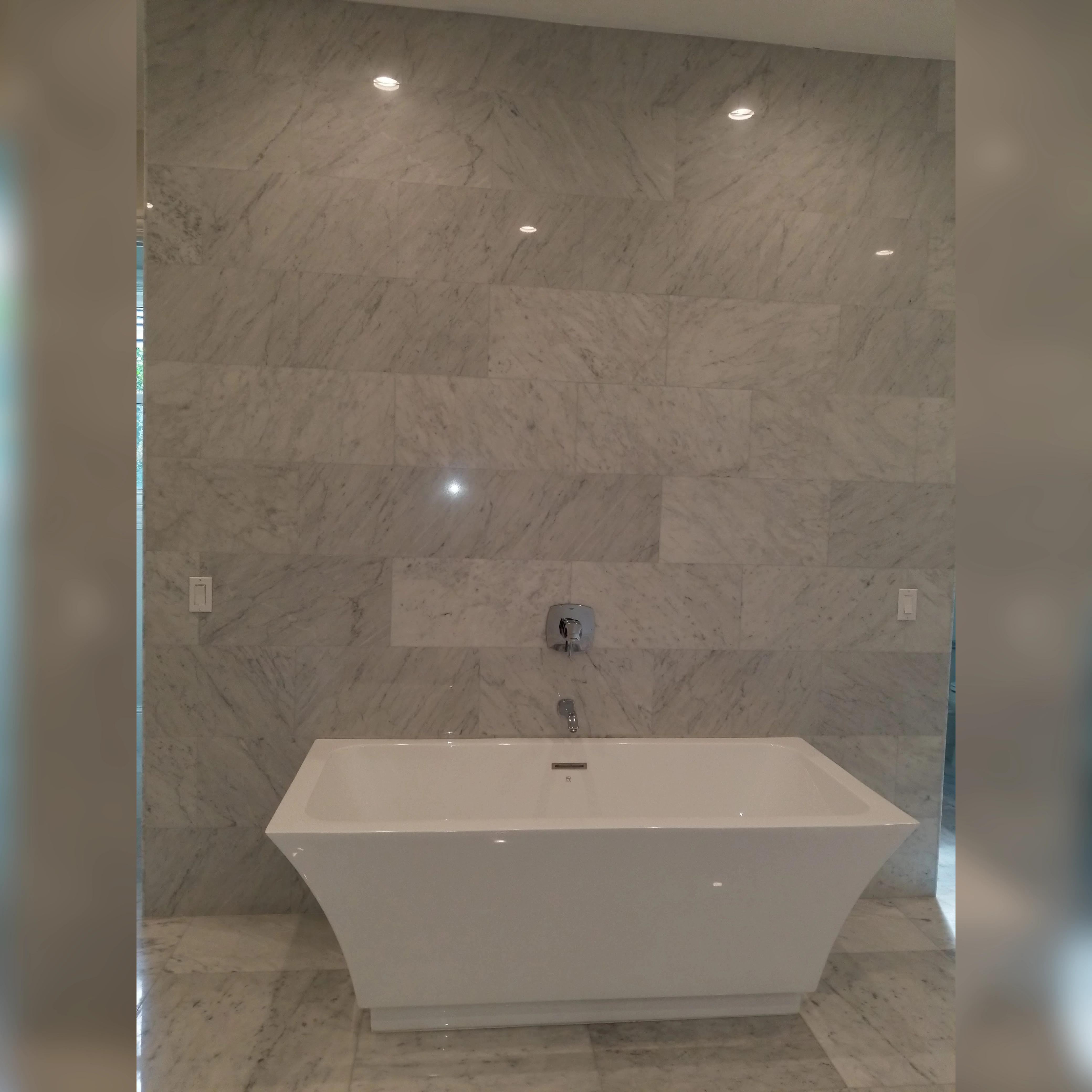 This bathroom features a sleek installation of Carrera marble tiles behind a freestanding sleek tub, creating a timeless and elegant backdrop with its natural veining and polished finish.