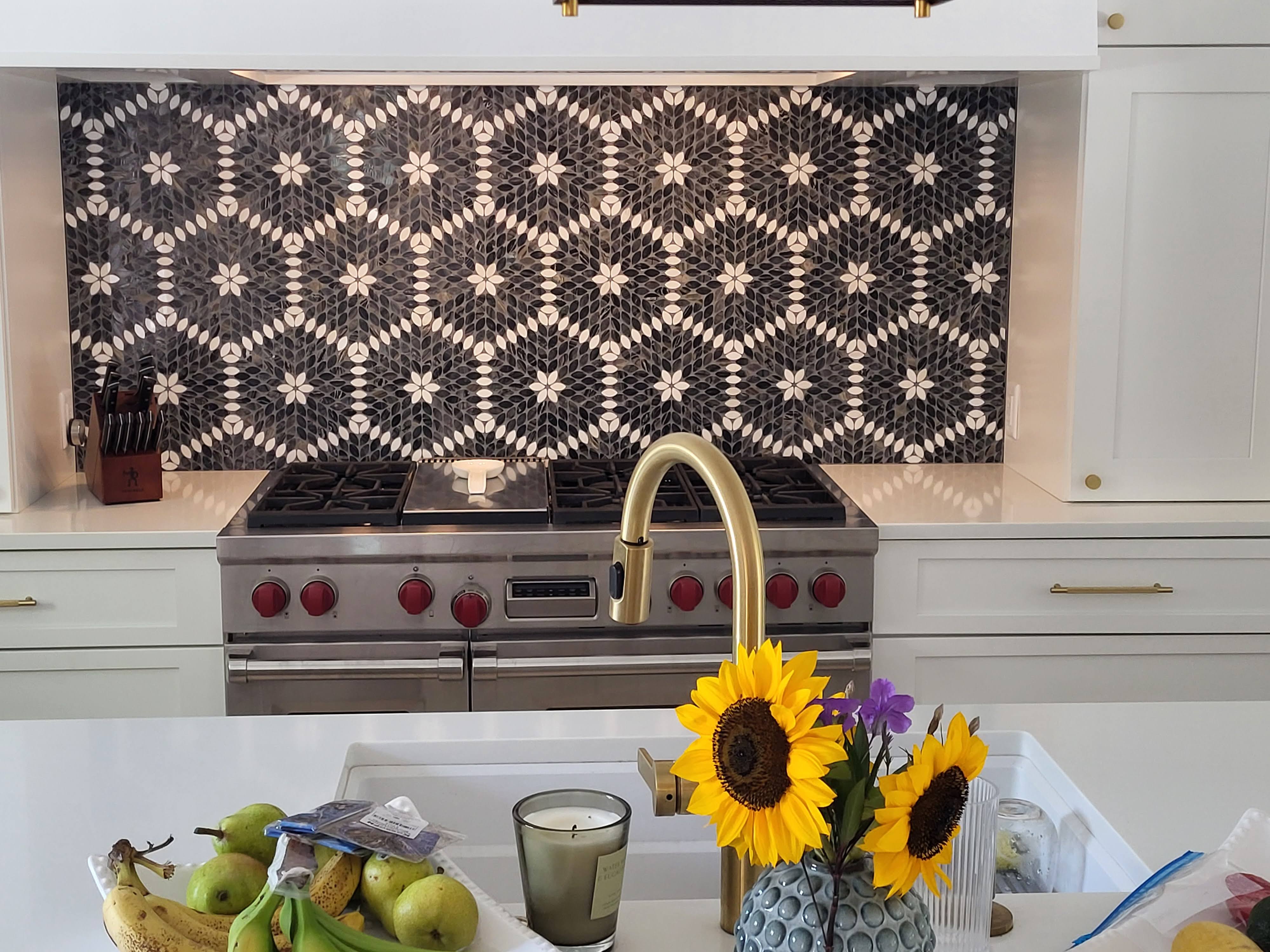 This kitchen backsplash features a striking mosaic tile design with intricate geometric patterns in contrasting black, white, and gray tones, serving as a bold focal point that beautifully complements the stainless steel appliances and crisp white cabinetry.