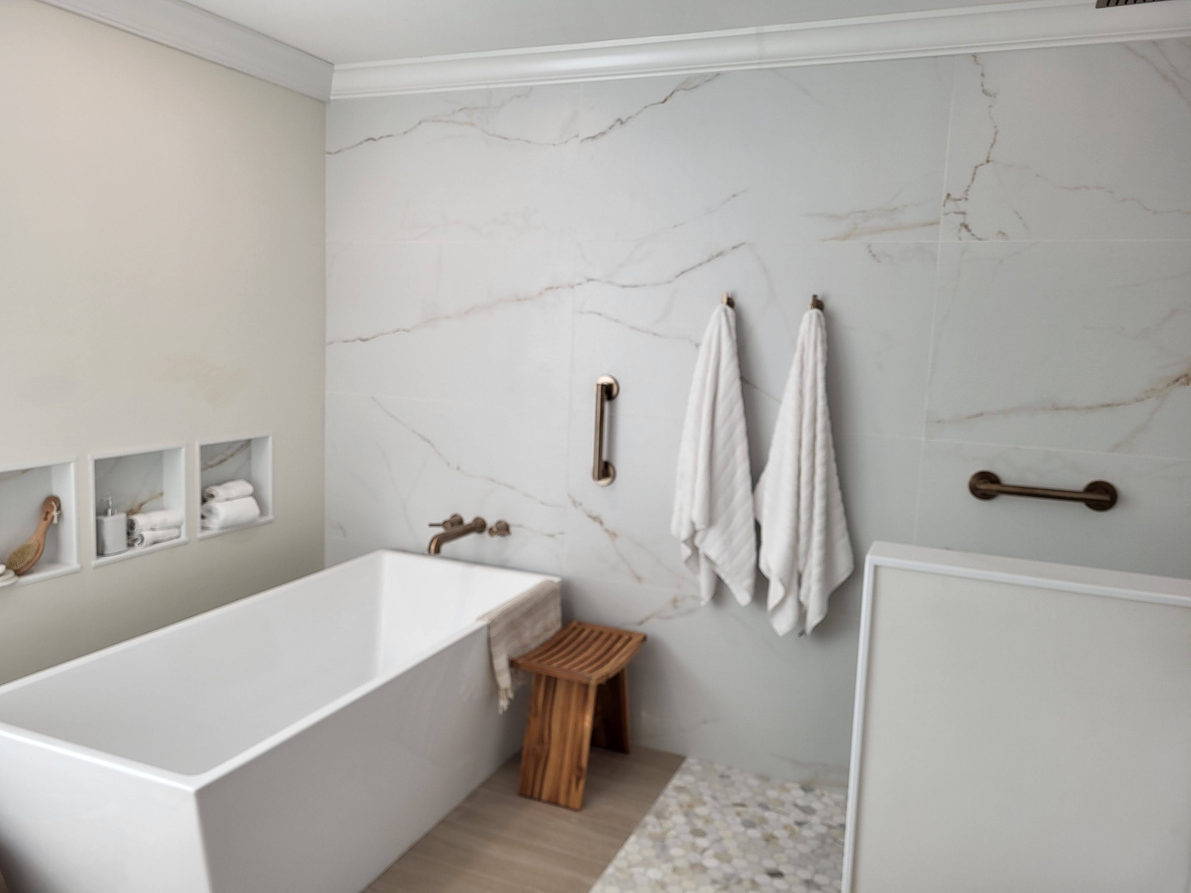 This contemporary bathroom features large-format porcelain tiles with soft, marble-like veining on the walls, a freestanding tub with brushed bronze fixtures, and a mosaic tile floor inset for added texture. Recessed niches provide convenient storage while maintaining a clean and minimalist design.