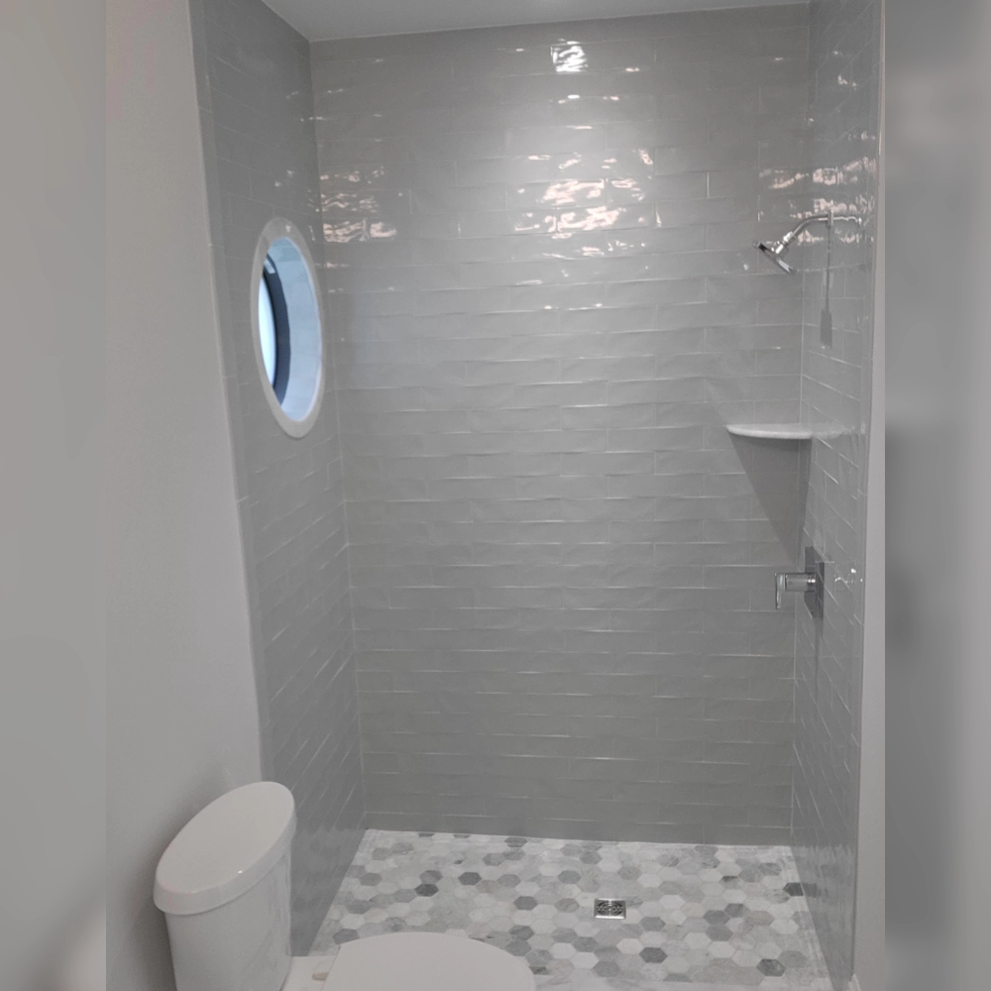 This modern shower features glossy, light gray subway tiles on the walls, paired with a hexagonal mosaic tile floor in complementary tones, creating a clean and contemporary design with a unique porthole-style window for added character.