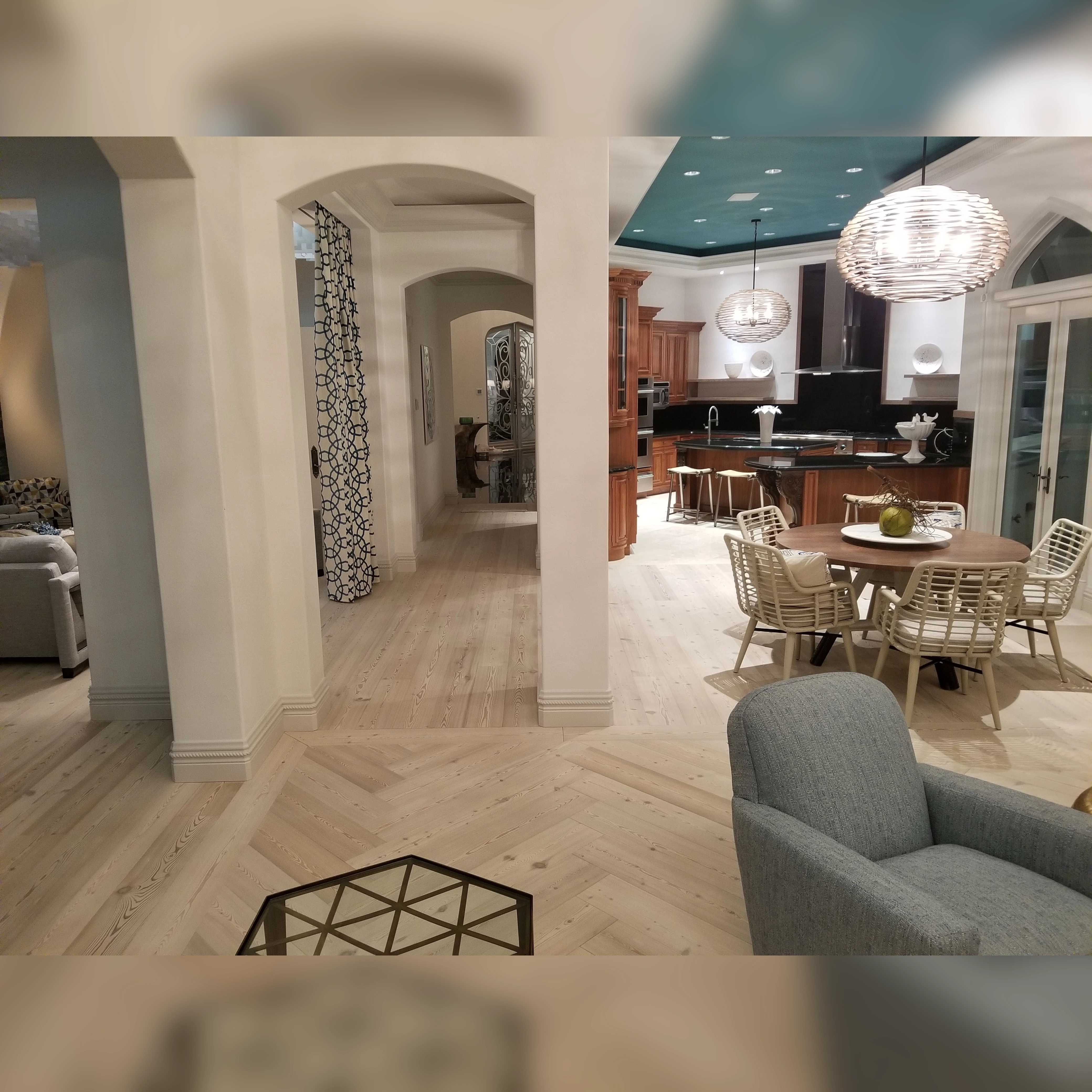 This open space features light wood flooring with a herringbone pattern in the living area, transitioning to straight planks in the kitchen and dining areas, highlighted by a border framing the hallway and bar room.