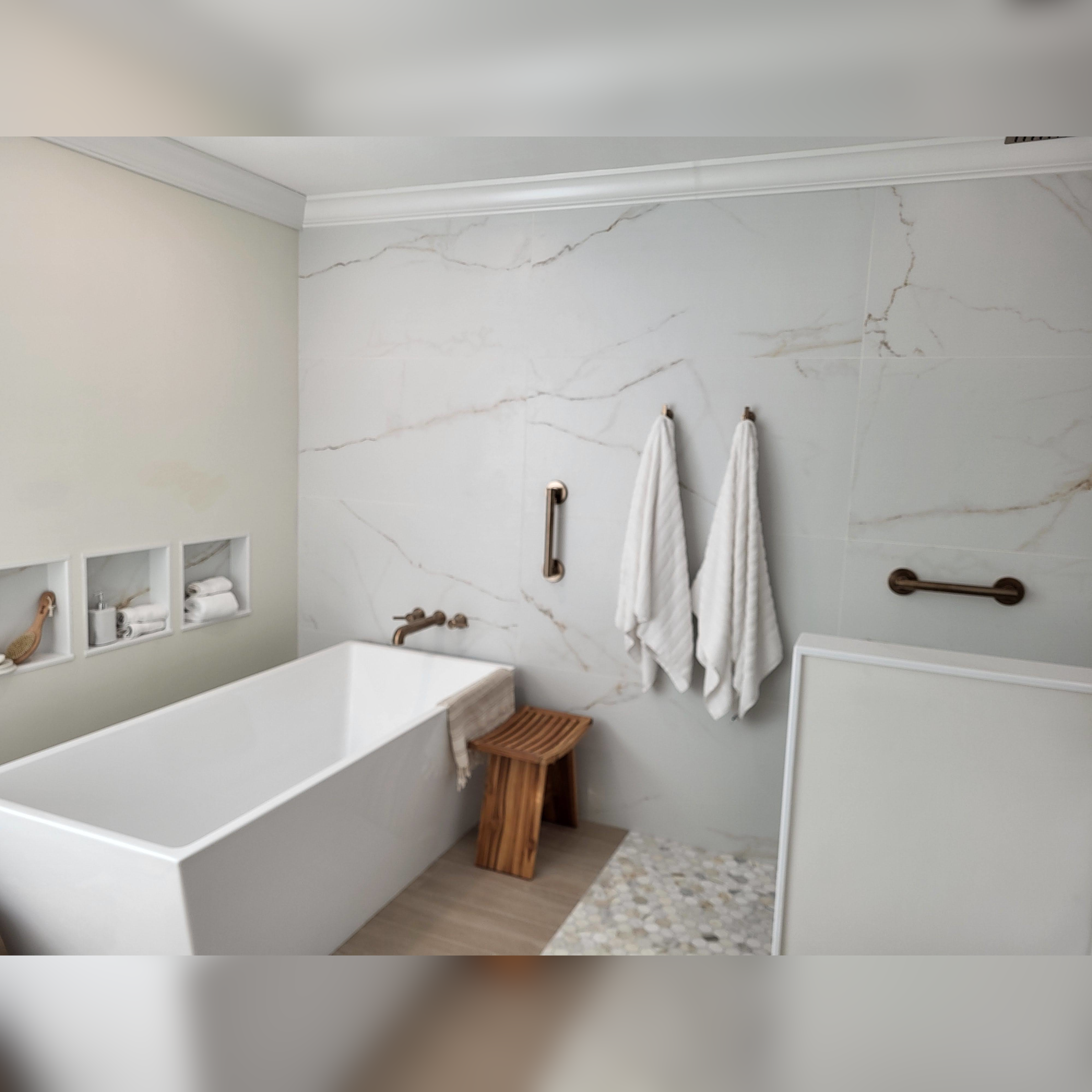 This contemporary bathroom features large-format porcelain tiles with soft, marble-like veining on the walls, a freestanding tub with brushed bronze fixtures, and a mosaic tile floor inset for added texture. Recessed niches provide convenient storage while maintaining a clean and minimalist design.