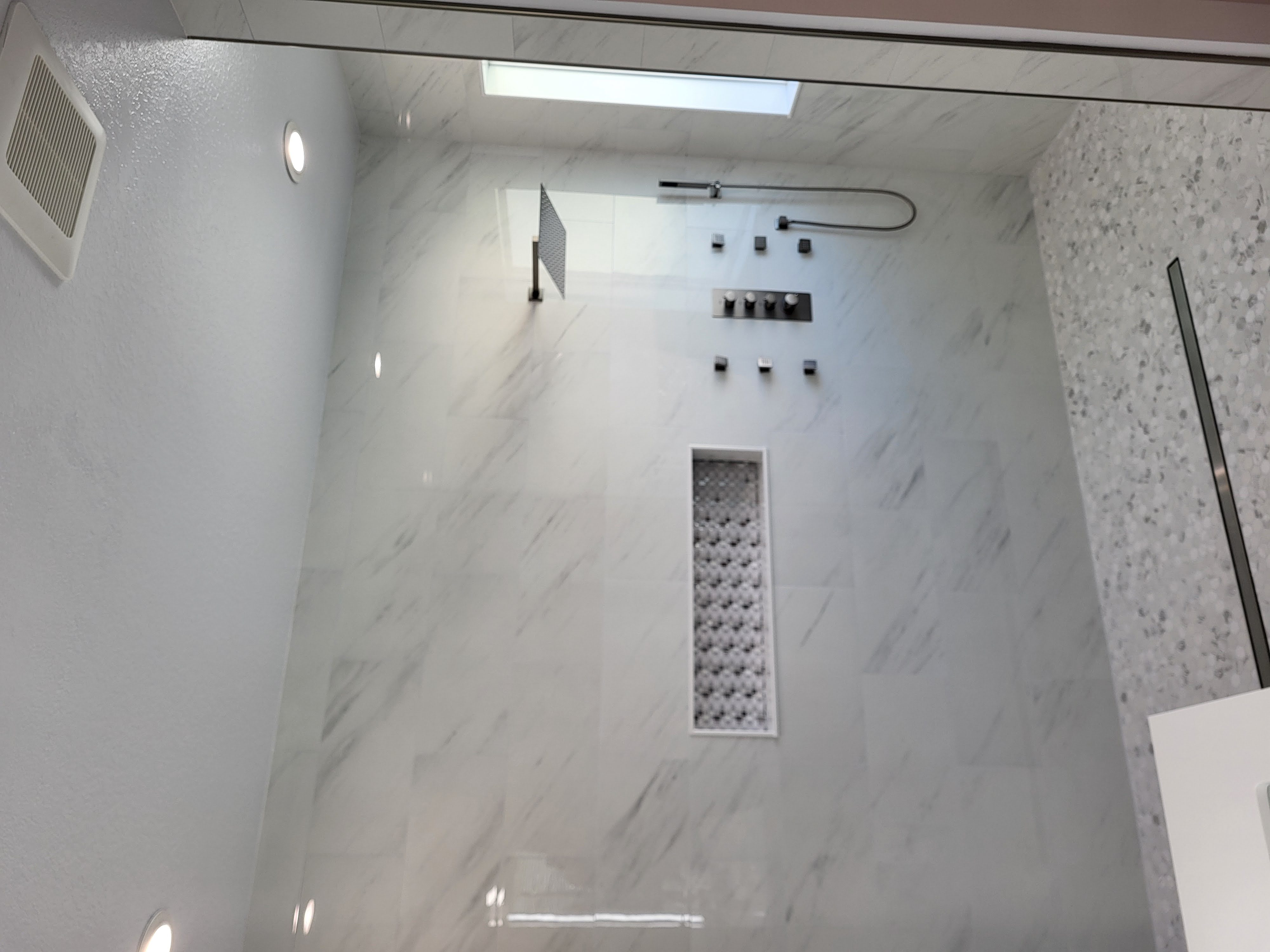 This modern shower features sleek, large-format porcelain tiles with subtle marble veining on the walls, paired with a mosaic tile floor for texture and contrast. A recessed niche with intricate mosaic detailing with rainfall shower head and body jets creating a spa-like experience.