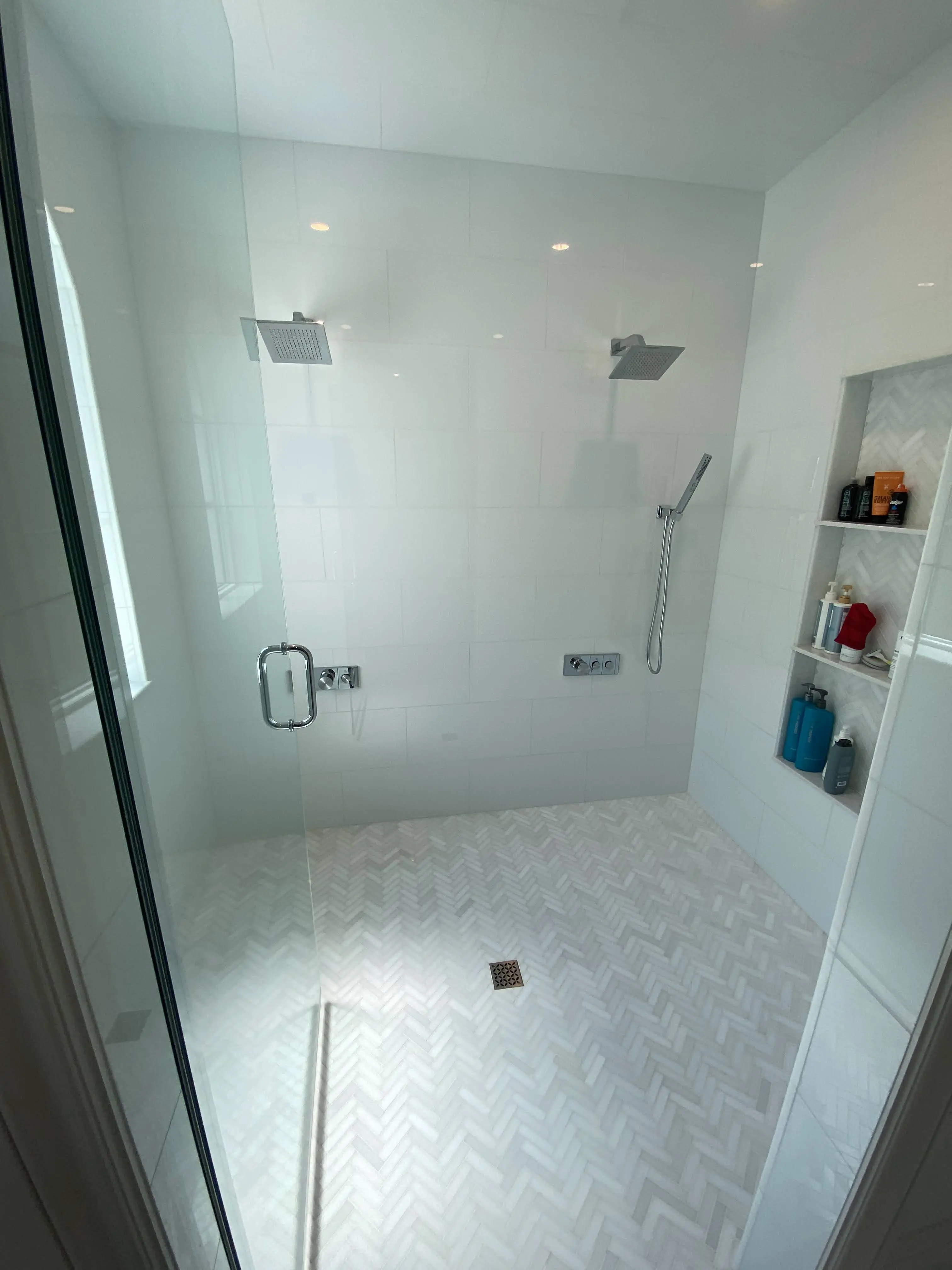 This spacious double shower features sleek porcelain Glassos tiles on the walls, paired with a white herringbone-patterned floor for added texture. Dual rainfall showerheads and a recessed niche provide both luxury and practicality in this modern design.