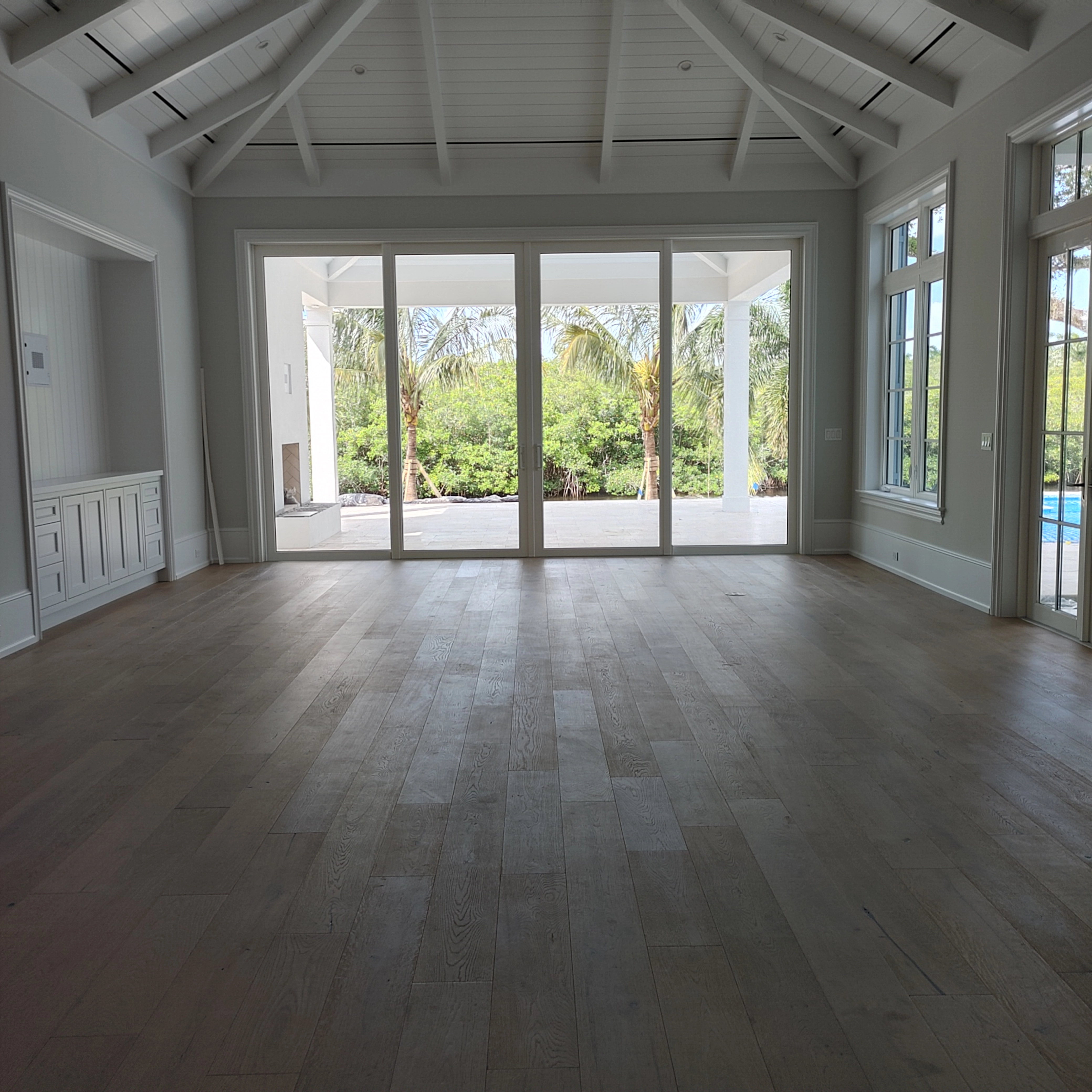 Wide-plank engineered hardwood flooring with a light oak finish creates a warm, natural foundation that complements the high-vaulted ceiling and enhances the airy, coastal feel of the space.