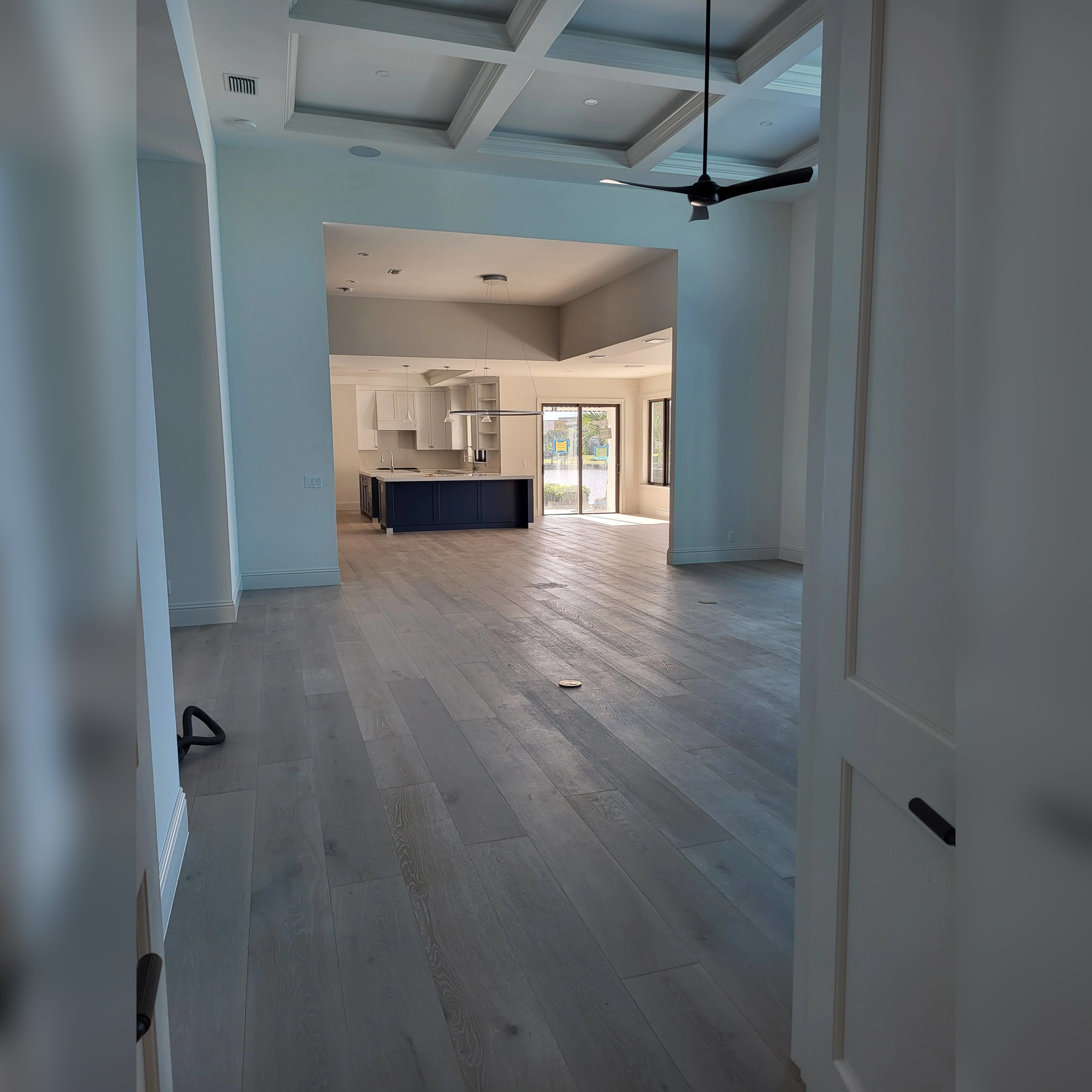 This space features engineered wood flooring in wide planks with a soft, gray-toned finish, creating a modern and cohesive flow between the living and kitchen areas, while complementing the coffered ceiling and clean architectural lines.